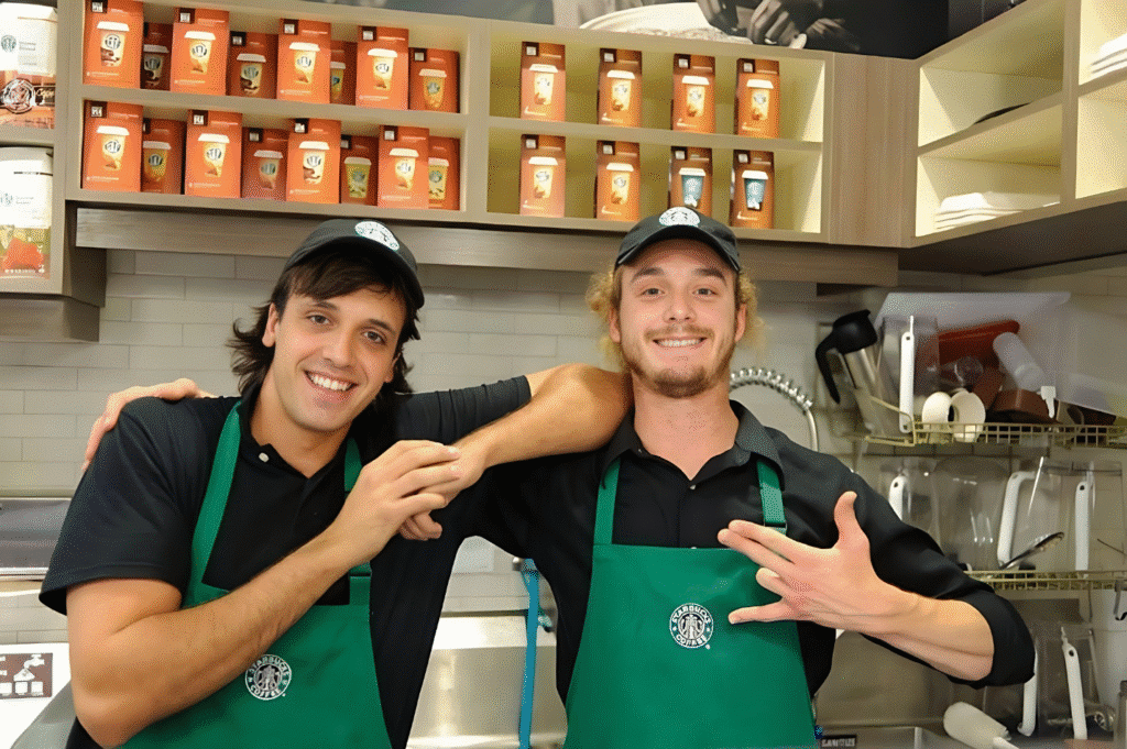 Two men in aprons smile for a photo inside a Starbucks, showcasing a friendly coffee shop atmosphere.