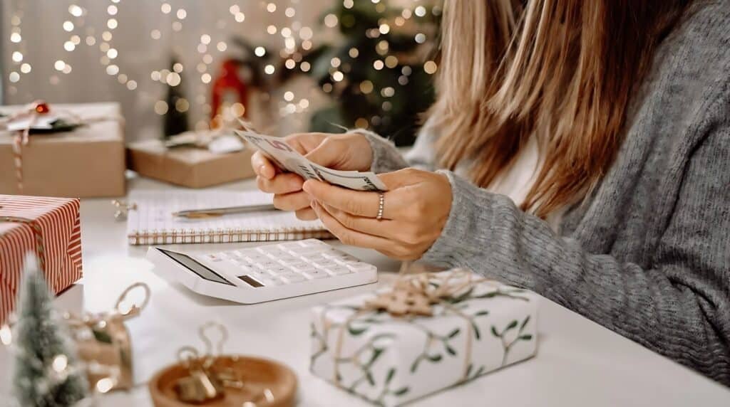 A woman holding a stack of money in her hands making budget for christmas while standing indoors near a decorated Christmas tree, with a table visible in the background.