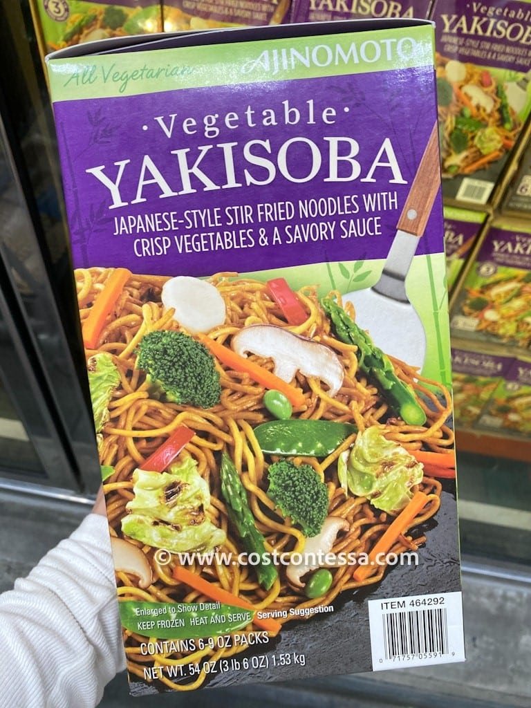 A hand holding a box labeled "Ajinomoto Yakisoba - Vegetable" featuring Japanese-style stir-fried noodles with crisp vegetables and a savory sauce, along with text indicating it contains multiple packs and serving suggestions. The box is designed to show details and suggests to keep frozen until heated for serving.