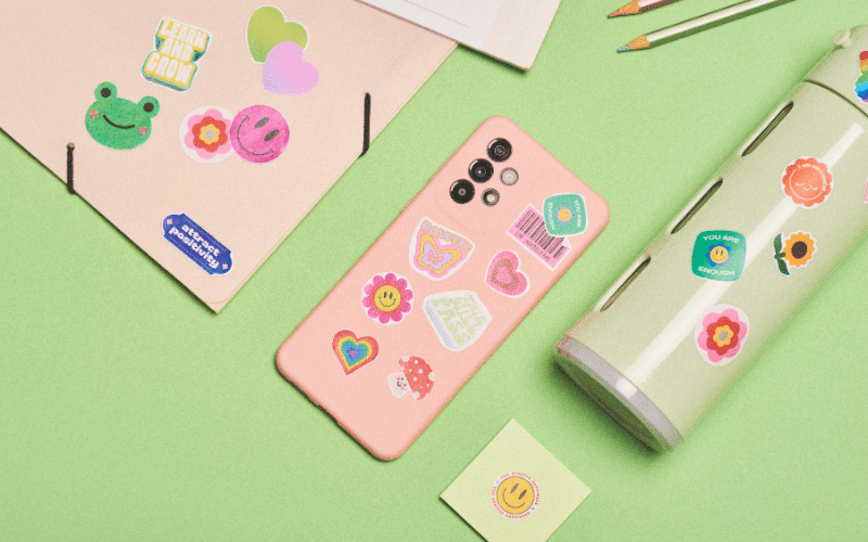 A pink phone case adorned with various stickers placed next to a pen, all set on a green surface.