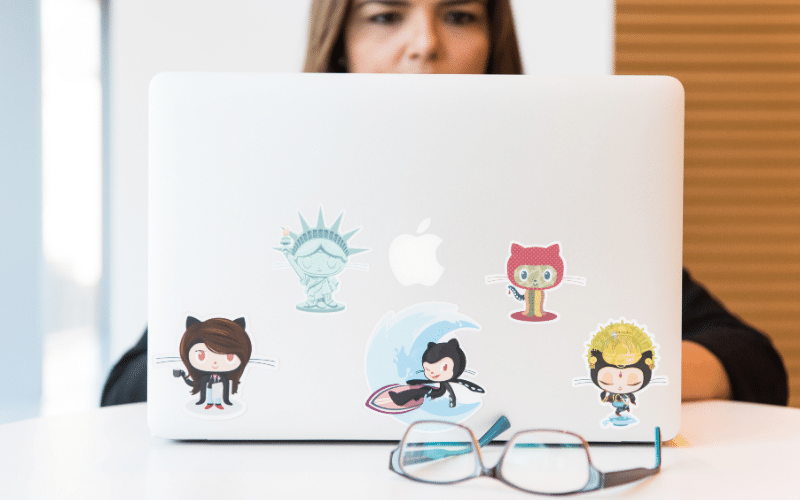 A laptop covered in various cartoon stickers, featuring playful designs including a smiling human face, sitting on a desk indoors.