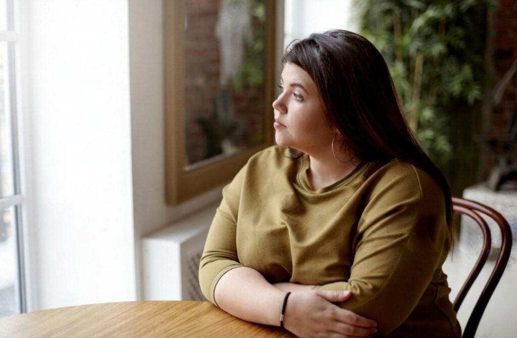 A woman sitting at a table, gazing out the window, reflecting on the challenges of side hustles like "$10K in 30 Days" coaching.