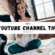 Smiling female YouTuber sitting on her bed with a camera and laptop, banner text saying Top 8 YouTube Channel Tips For Beginners.