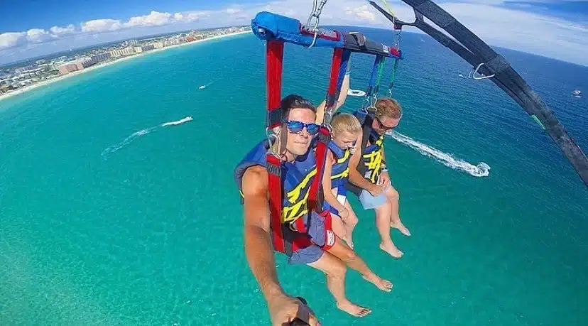 A family enjoys a parasail ride over the ocean in Destin, Florida, with vibrant blue waters and a clear sky in the background.