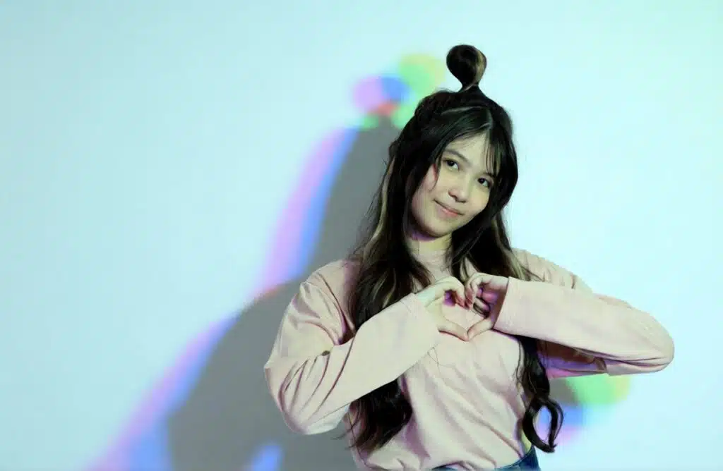 Girl in a pink long-sleeve top makes a heart with her hands in front of a white wall with colorful shadow lighting illustrating Content Ideas for TikTok KPop Fanpage