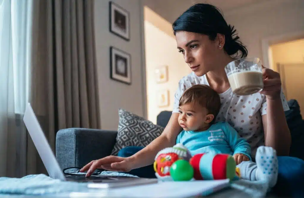 Mom working on a laptop with baby on her lap, holding a mug at home—mom blogger lifestyle