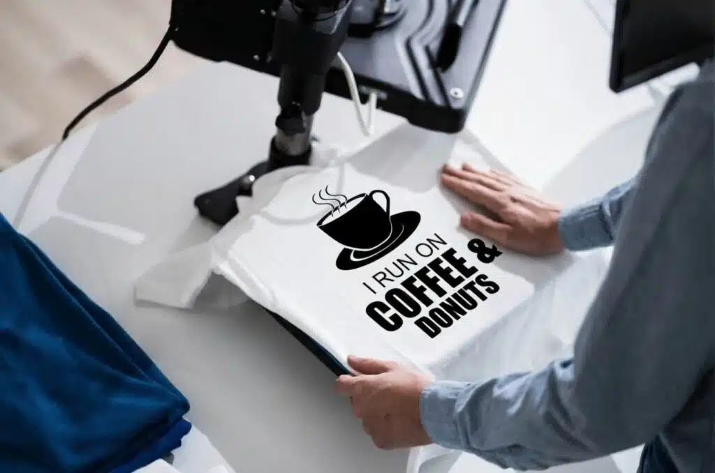 A person using a heat-press machine to transfer a “I Run on Coffee & Donuts” design onto a white T-shirt, illustrating print-on-demand as one of the Good Side Hustles for Women.