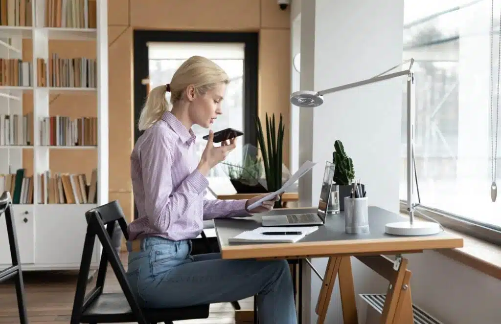 A woman working at a desk in a bright home office, speaking into her phone while reviewing documents and using a laptop, representing Good Side Hustles for Women such as virtual assistant work.