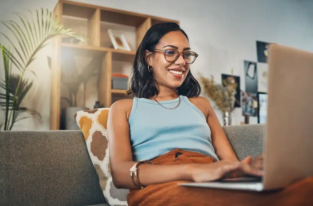 A smiling woman sitting on a couch using a laptop at home, representing blogging as one of the Good Side Hustles for Women.