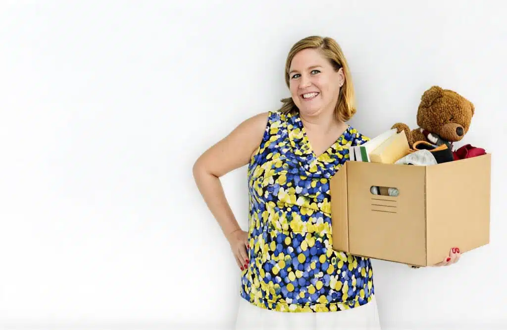 A smiling woman holding a cardboard box filled with items like books, clothing, and a teddy bear, representing decluttering and selling unused belongings as one of the Good Side Hustles for Women.