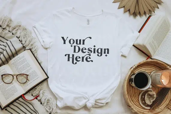 Minimal white t-shirt mockup with text reading “Your Design Here,” styled with books and coffee, representing how to sell t-shirts online using simple text-based designs that convert without complex graphics.